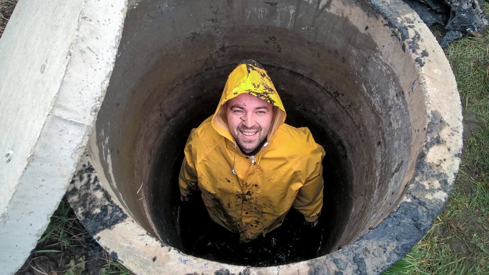 A man wearing a yellow raincoat stands inside a concrete well, smiling up at the camera. Dirt and mud are visible on his coat and around the well, illustrating the hands-on dedication of Cesspool Services Long Island.