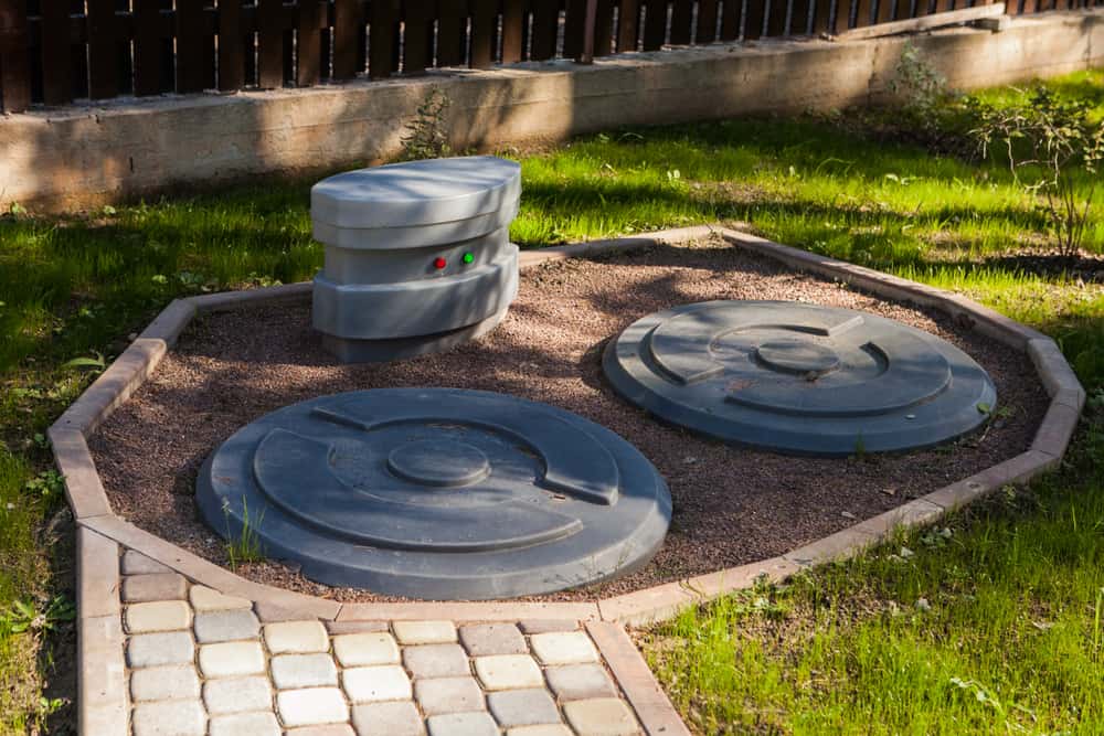 Two large circular septic tank covers and a smaller vent cover are placed in a gravel area, edged with stone bricks and surrounded by grass. Regular septic tank maintenance Long Island residents trust is essential for maintaining this setup.