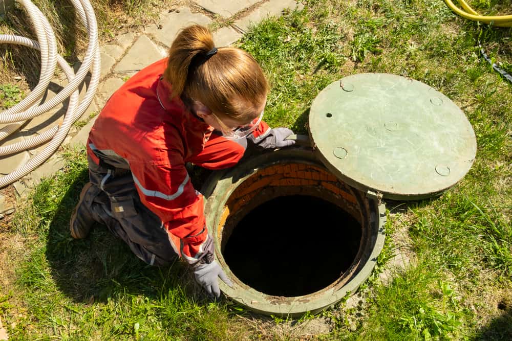 A person in protective gear and gloves opens a circular outdoor manhole cover, peering inside, with hoses around on the grass—typical of septic tank maintenance Long Island.