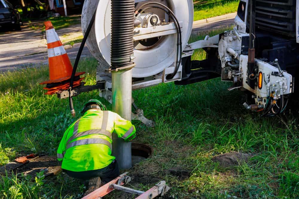 A worker in a bright yellow jacket operates machinery to clean a sewer drain, with an orange traffic cone visible on the equipment, showcasing the expertise of Cesspool Services Long Island.