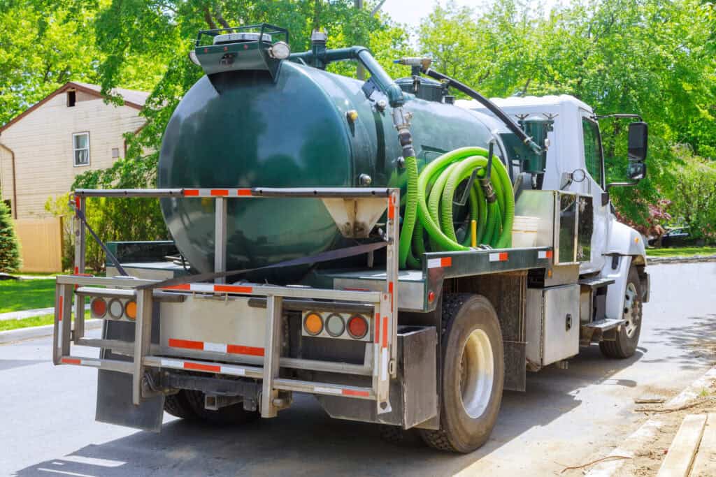 A white and green septic truck, essential for Cesspool Services Long Island, is parked on a residential street. It features a large green tank and hoses, with a house and trees in the background.