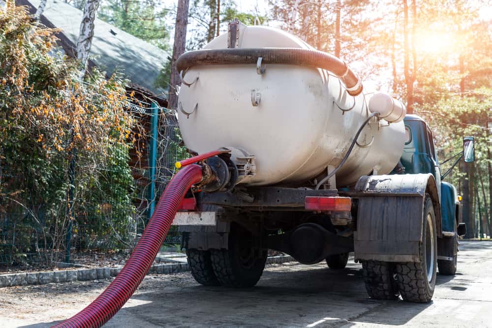 A septic tank truck with a large tank and hose is parked on a forested property, with sunlight filtering through the trees in the background, providing top-notch Septic Tank Services Long Island residents can rely on.