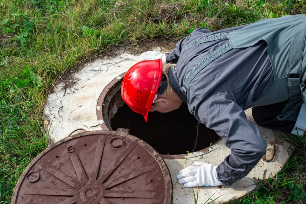 A person wearing a red helmet and gray overalls leans over to inspect an open manhole, likely checking for any septic tank issues. They are kneeling on grass with the removed manhole cover nearby, as part of their detailed inspection routine.