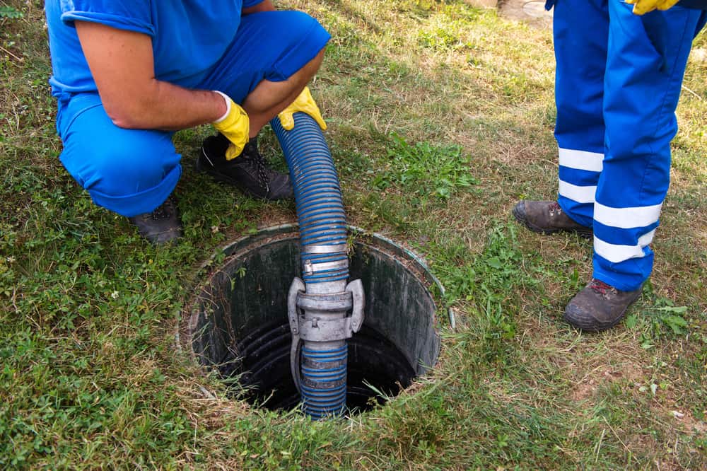 Two workers in blue uniforms and yellow gloves are handling a large hose inserted into an open manhole in a grassy area, providing expert septic tank services. One worker is crouching, while the other stands nearby, ensuring everything runs smoothly.