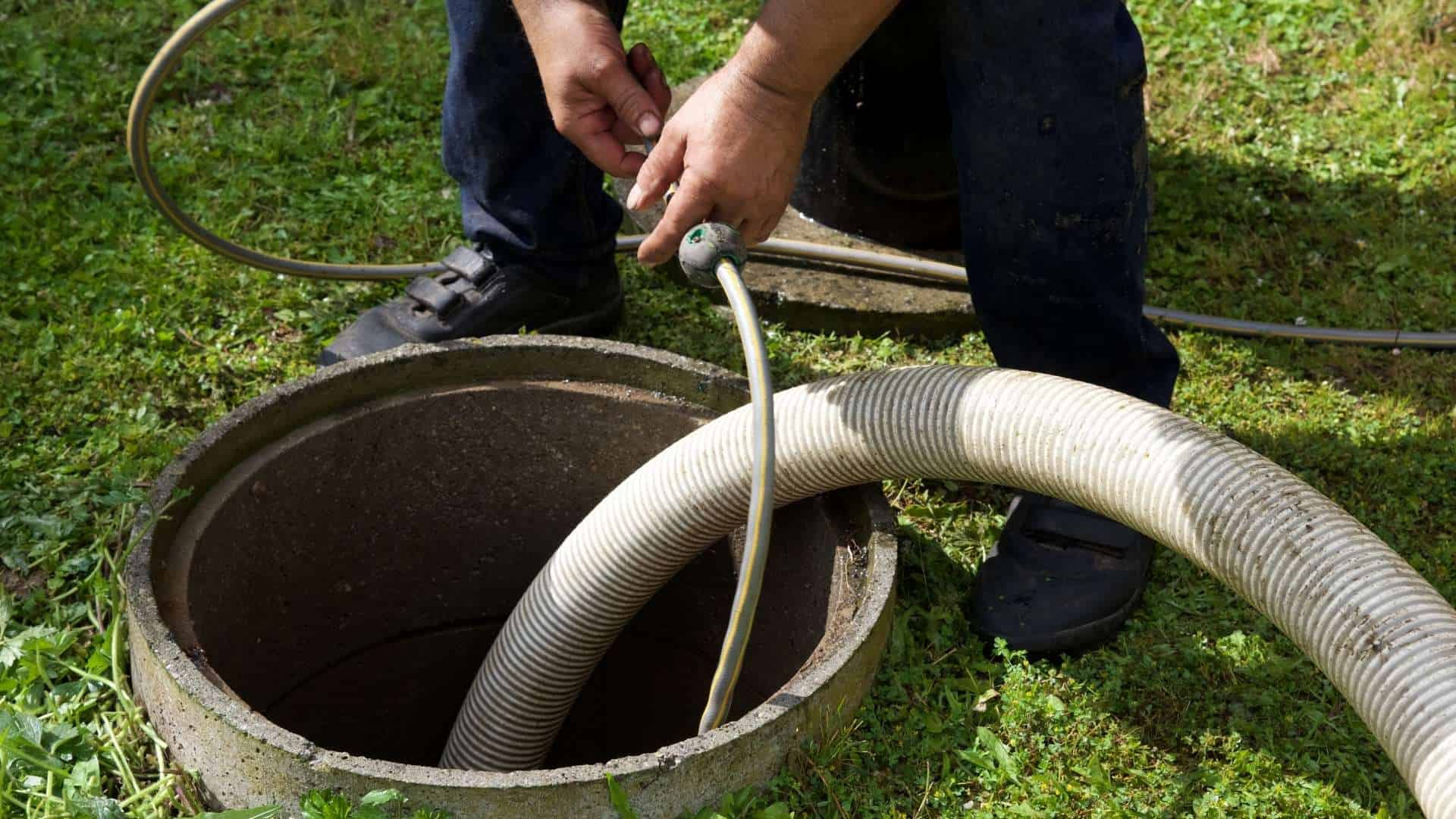 A person handling a hose attached to an open septic tank lid, surrounded by green grass. The individual is wearing dark pants and shoes, and is bending over to manage the equipment, indicating septic tank services or cleaning activity.