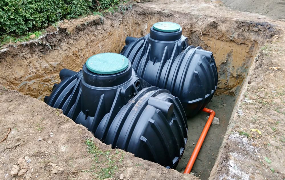 Two large black plastic septic tanks are partially installed in a rectangular underground pit, showcasing expert septic tank services. The tanks are connected by an orange pipe, with surrounding earth exposed to reveal layers of soil and grass.