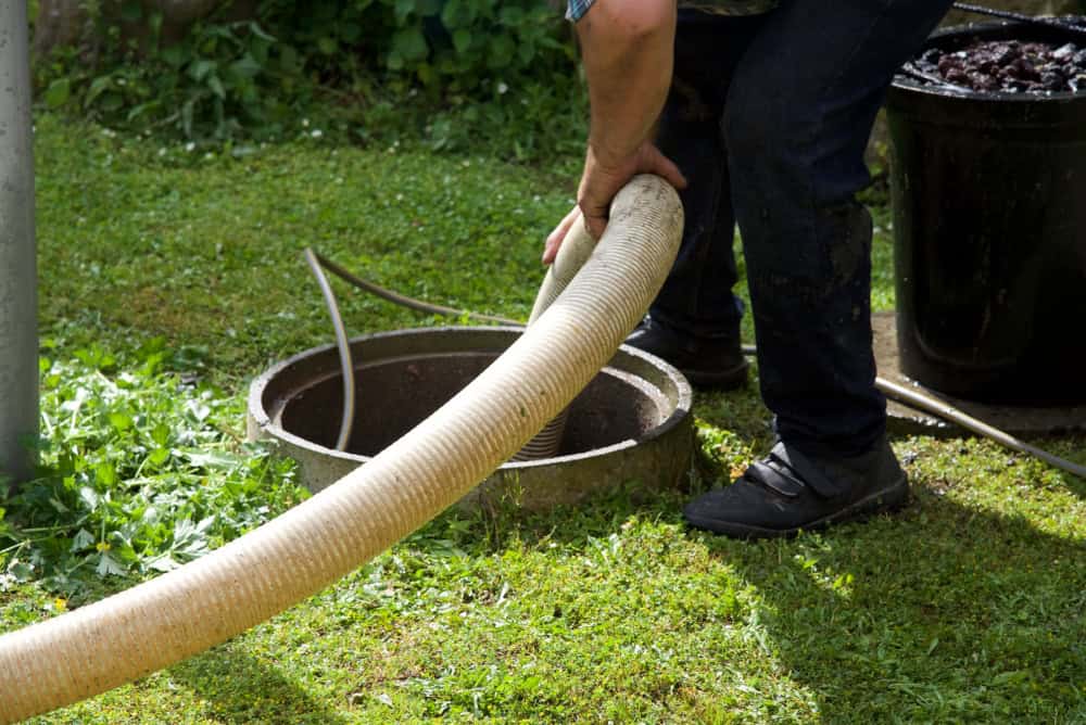 A person is holding a large hose over an open septic tank in a grassy area, showcasing expert Septic Tank Services. Nearby, another black container stands ready. The person is dressed in dark pants and shoes, efficiently managing the task at hand.