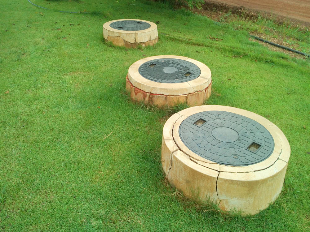 Three concrete manhole covers, possibly linked to septic tank services, are arranged in a row on a lush grassy area. Circular and slightly elevated with dark metal lids, they sit amid the well-maintained lawn, while a dirt path weaves through the background.