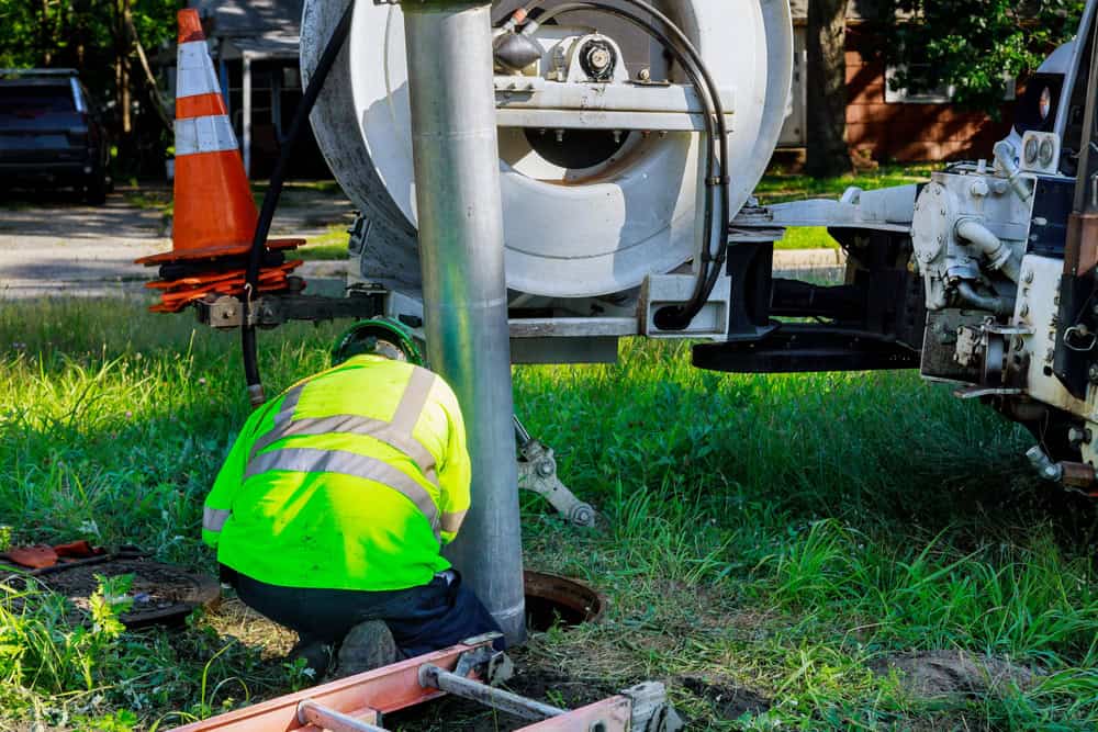 A worker in a fluorescent vest and hard hat kneels by a large vacuum truck, inspecting or cleaning a sewer line as part of essential septic tank services. An orange traffic cone is placed nearby, and a ladder lies on the grass beside the worker.