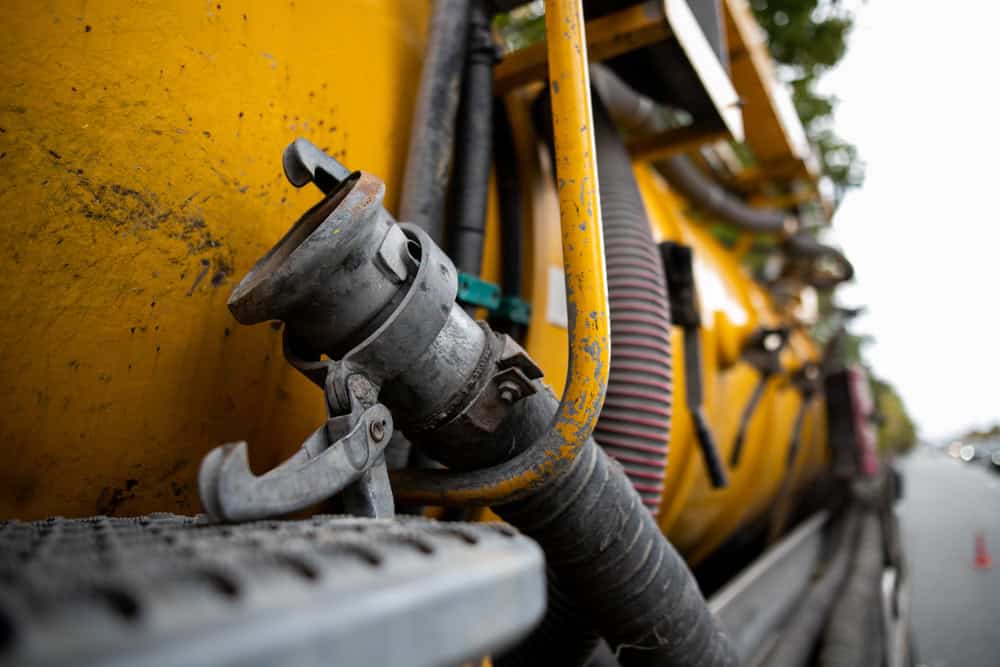 Close-up of industrial tubing and valve equipment attached to a large yellow tank on a vehicle, likely used for septic tank services or waste management. The image is taken from a low angle, focusing on the valve and metal structures.