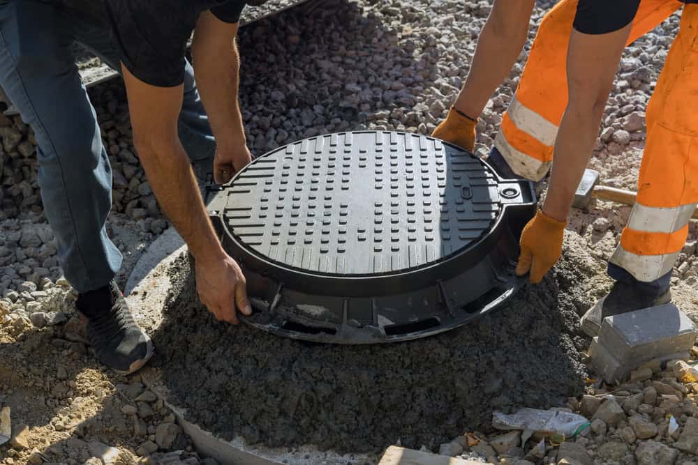 Two workers from a septic tank services team are installing a round manhole cover on the street. They are wearing protective gloves and clothing, including orange safety pants. The surrounding area consists of gravel, and the manhole is set in concrete.