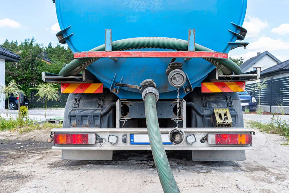 Rear view of a blue sewage tanker truck with hoses connected for waste disposal. The vehicle has a red and white license plate area, warning signs, and is parked on a dirt road near residential buildings.