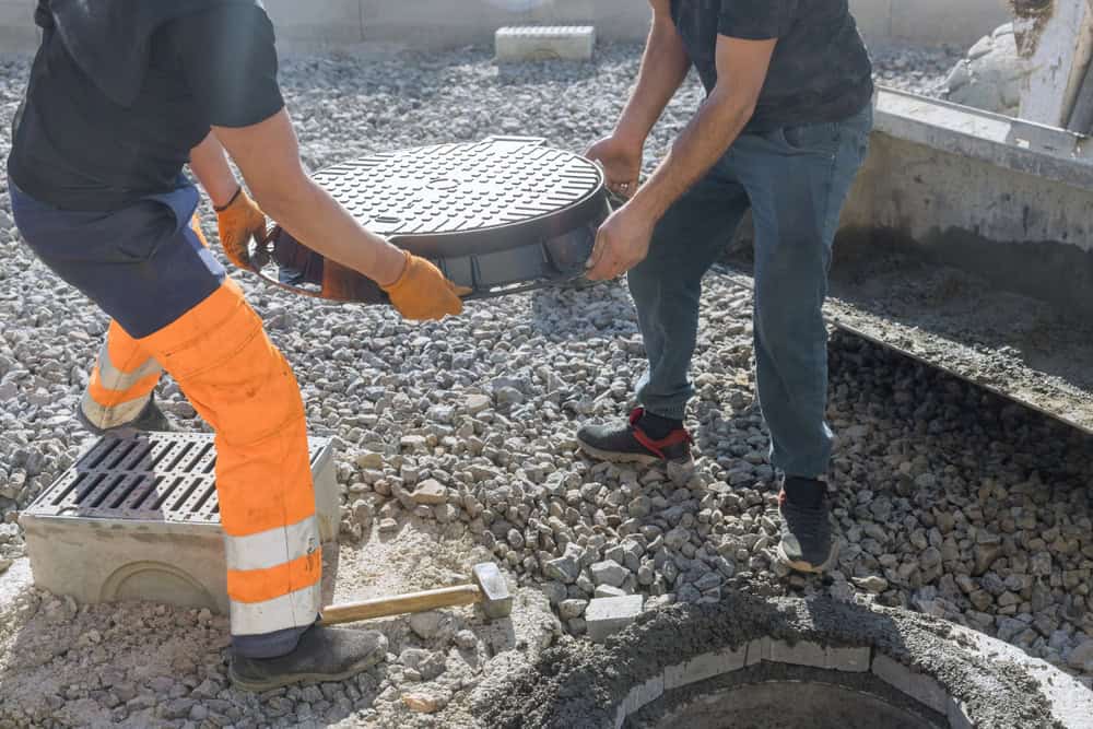 Two workers wearing gloves and protective clothing are lifting a manhole cover over a concrete opening at a construction site for septic tank services. The ground is covered in gravel, and a concrete frame is partially visible.