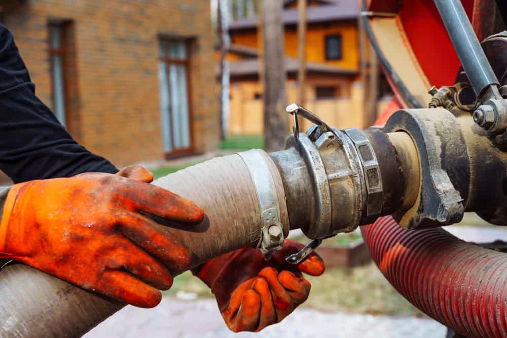 A person wearing orange gloves is connecting a large hose to a metal fitting outdoors, likely for septic tank services. The background showcases a brick building and a wooden house, enveloped by trees and grass.