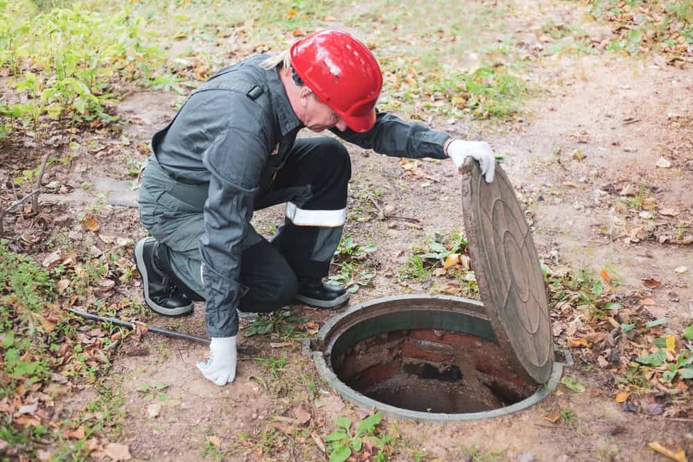 A worker in a red hard hat and gray coveralls kneels beside an open manhole, inspecting it with the precision of septic tank services. The area is surrounded by dry leaves and grass.