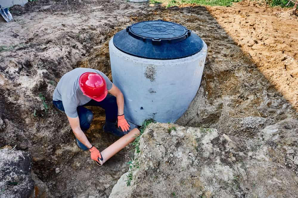 A worker in a red hard hat and orange gloves installs a pipe into the ground beside a large, cylindrical concrete septic tank. The surrounding soil suggests recent excavation work typical of expert septic tank services.