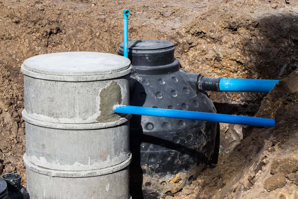 A black septic tank with blue PVC pipes is partially buried in the ground next to a concrete well chamber, illustrating a typical project by professional septic tank services. The surrounding area is dug out, clearly revealing the installation in progress.