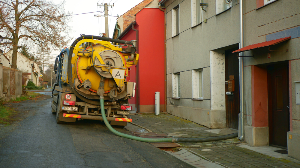 A large yellow septic truck, marked with "Cesspool Services," is parked on a narrow street, its hoses snaking into a building on the right. The closely situated houses suggest a residential area. In the background, you can see a leafless tree and telephone poles stretching skyward.