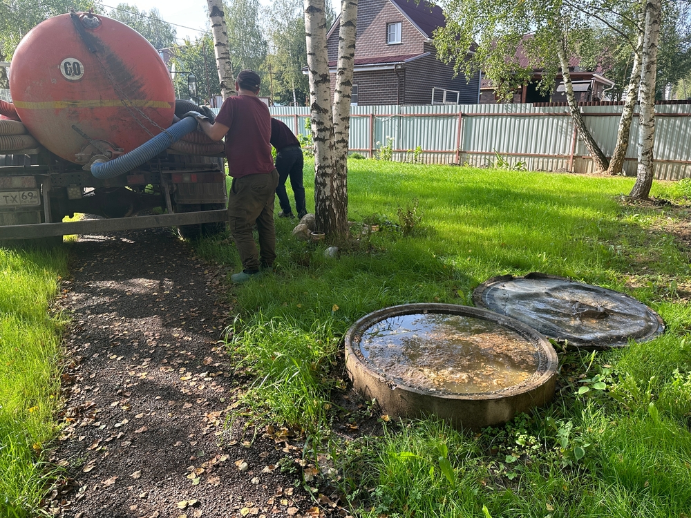 Two workers are standing near an open septic tank in a grassy area, providing septic tank services. One operates a hose connected to a large tanker truck. In the background, there are trees and a residential building, illustrating the serene yet essential operation of cesspool maintenance.