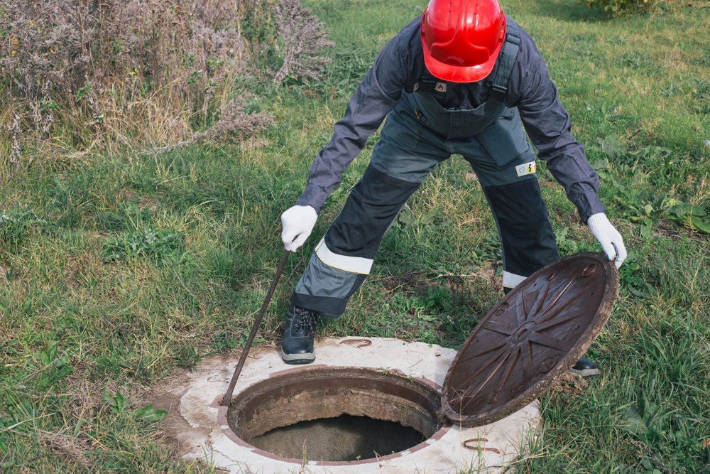 A worker wearing a red helmet and protective clothing lifts the circular metal cover of a manhole in a grassy area, using a tool, as part of septic tank services.