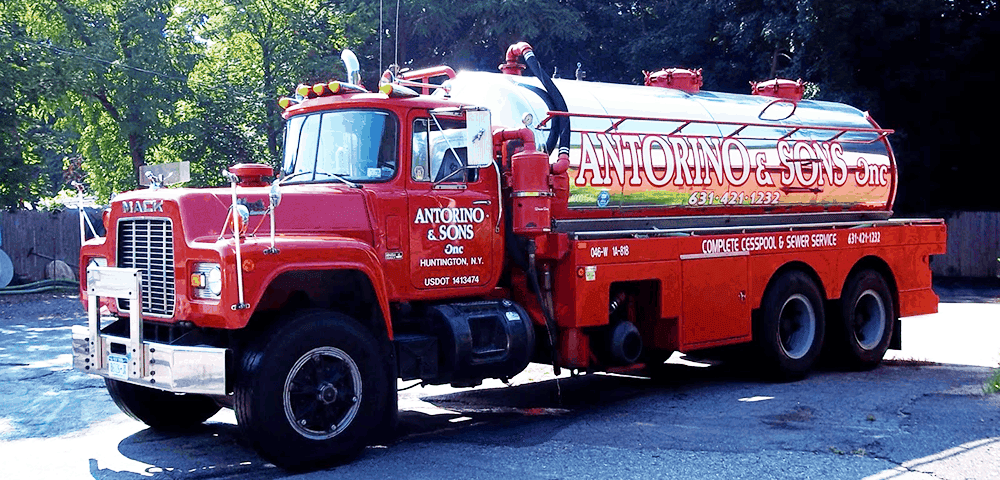 A bright red tanker truck with "Antorino & Sons" lettering gleams under the sun, nestled among trees and a wooden fence. With chrome details and a large tank, it's poised for commercial or septic tank services.