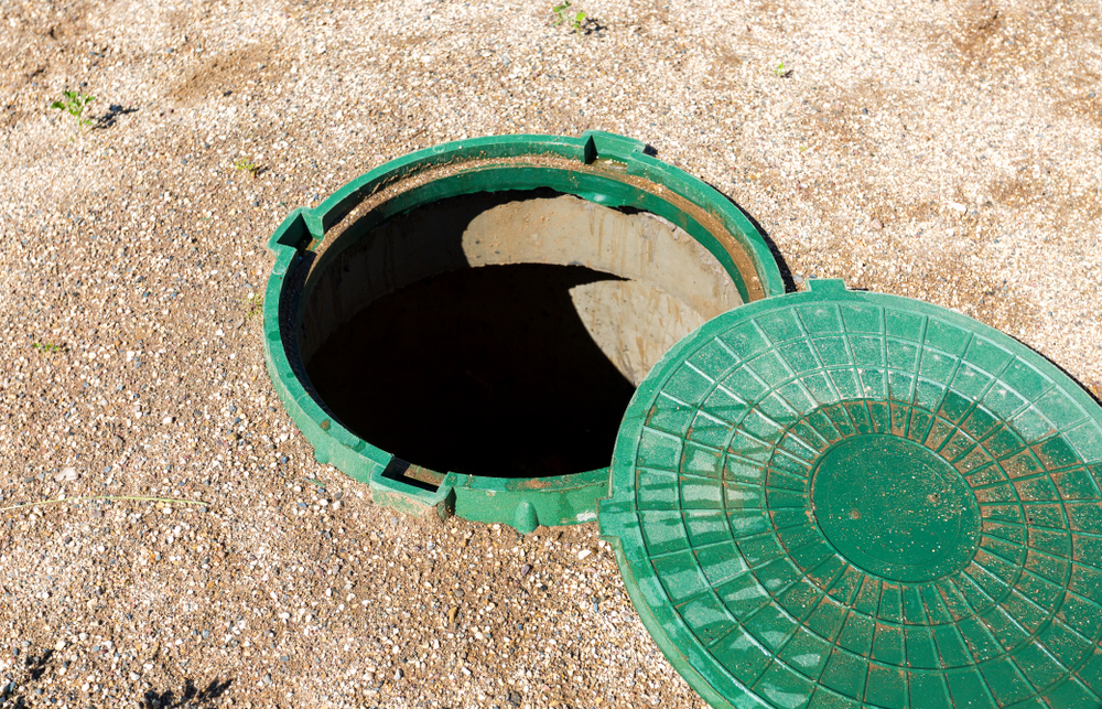 A green manhole cover is partially open, revealing the dark opening of a sewer or utility access point. The cover rests on a gravelly ground surface, with some small plants visible.