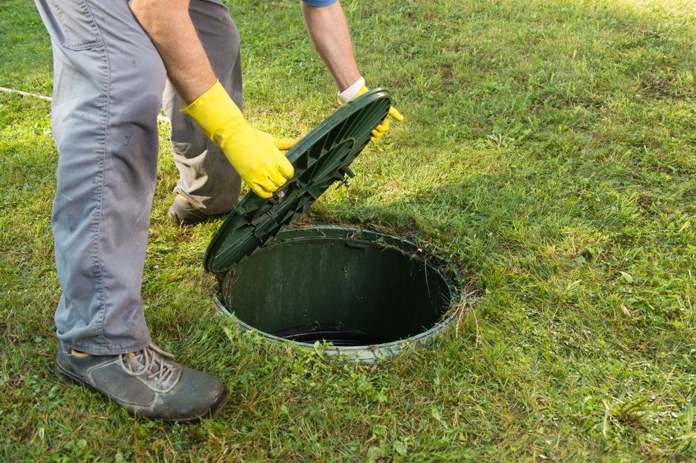 A person wearing yellow gloves and gray pants lifts the lid of a green septic tank embedded in a grassy lawn. The lid is partially open, revealing the circular opening of the tank.