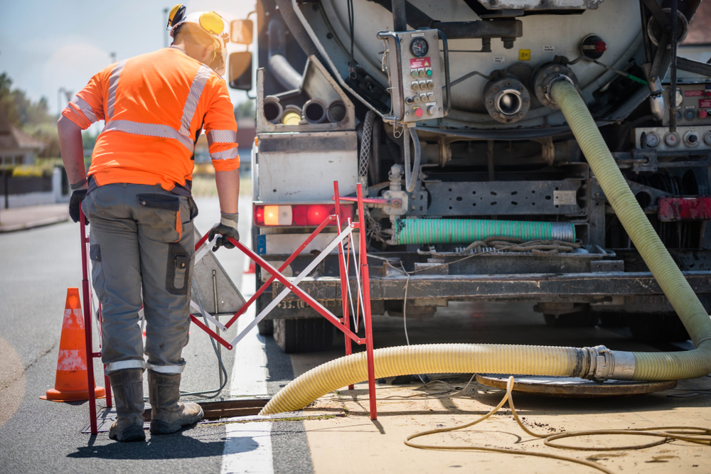 A worker in high-visibility gear operates machinery next to a large truck on a road. Hoses are attached to the truck, and traffic cones are placed nearby. The scene suggests road maintenance or construction work in progress under daylight.