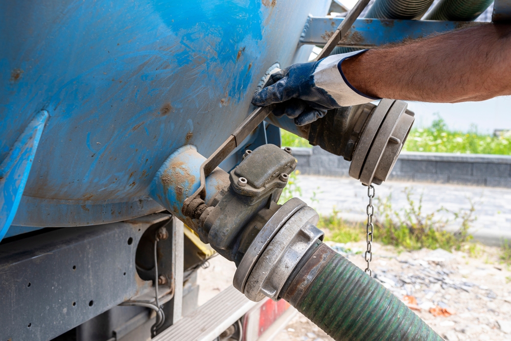 A person's gloved hand tightens a bolt on a large blue tank with a wrench. A green hose is attached to a valve on the tank. The background shows a stone wall and greenery.