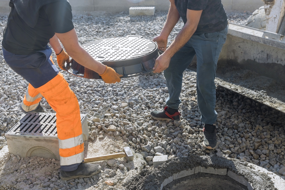 Two workers in safety gear install a round manhole cover onto a concrete opening at a construction site, surrounded by gravel and tools. One is wearing orange and black, the other blue and black.