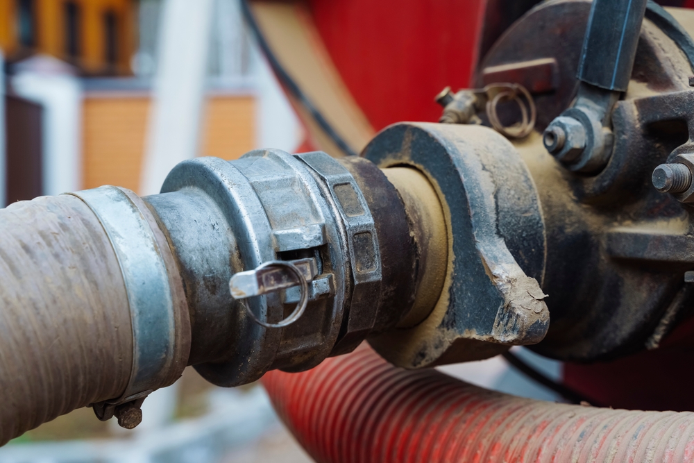 Close-up of a metal clamp securing a large hose to a pipe connection on industrial machinery. The equipment shows signs of wear and is coated with dust. The background is slightly blurred, hinting at an outdoor setting.