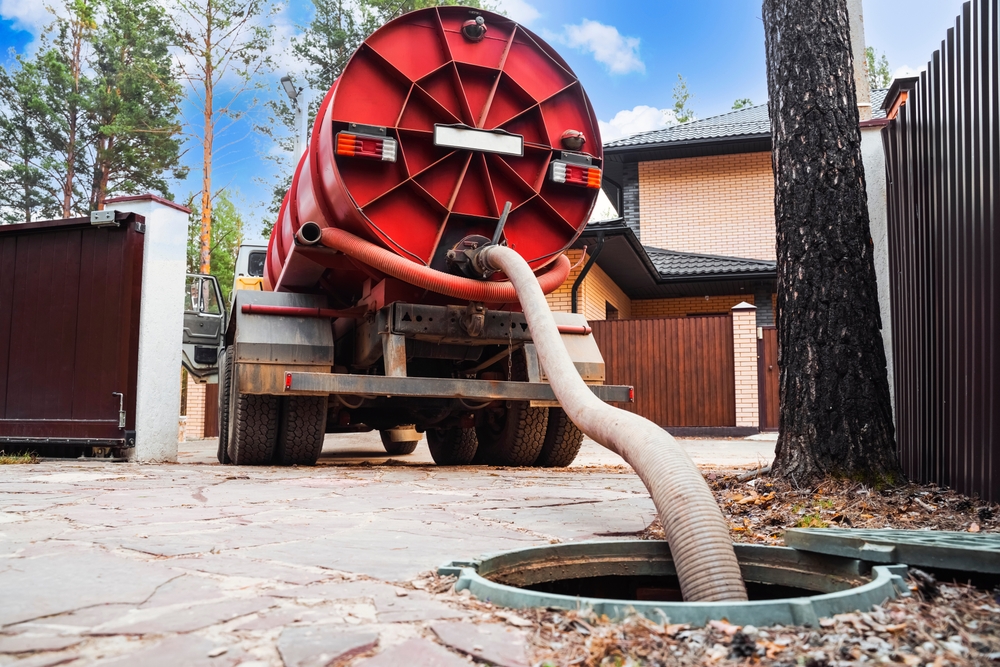 A septic tank truck with a large red cylindrical tank is parked in a residential area. It has a hose inserted into an open septic tank, performing maintenance. Trees and a brick house are in the background.