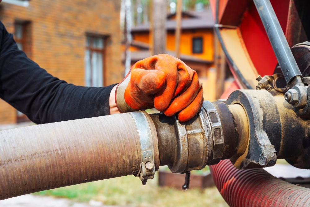 A person wearing orange gloves is connecting a hose pipe to a large metal fitting outdoors. In the background, there are trees and brown brick buildings. The scene suggests a construction or maintenance work environment.