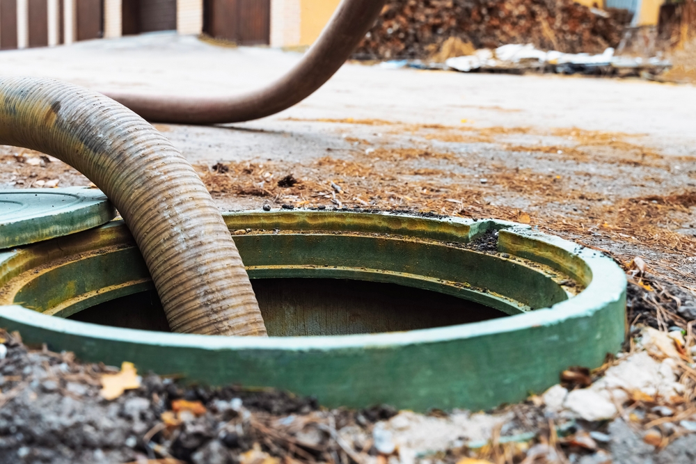 A large hose is inserted into an open sewer manhole on the ground. The image shows a close-up of the green metal cover and surrounding dirt. A pile of leaves and a few structures can be seen in the blurred background.