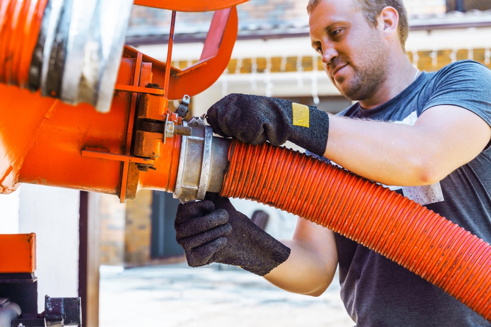 A man wearing black gloves is working with a large orange hose, securing it to a metal coupling. He is outdoors, focused on the task. The background features a blurred brick building and a slightly open garage door.