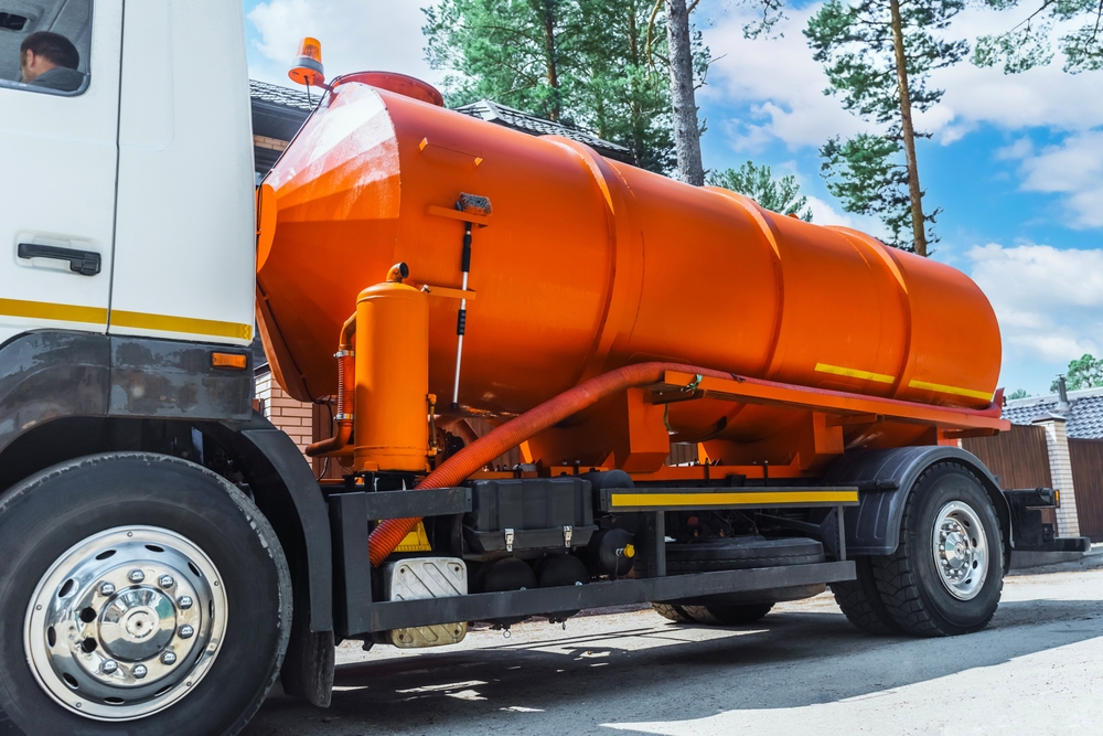 Orange vacuum truck with a large cylindrical tank parked on a street. The vehicle is designed for cleaning and waste removal. It is positioned next to a building, with trees and a blue sky in the background.