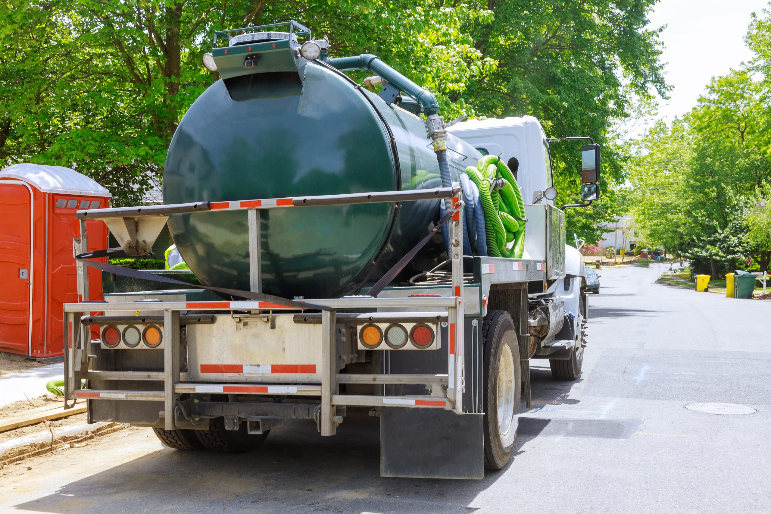 A septic tank maintenance truck on a suburban street, with a large green tank and hoses attached. There are trees, a portable toilet, and trash bins in the background under a clear sky.